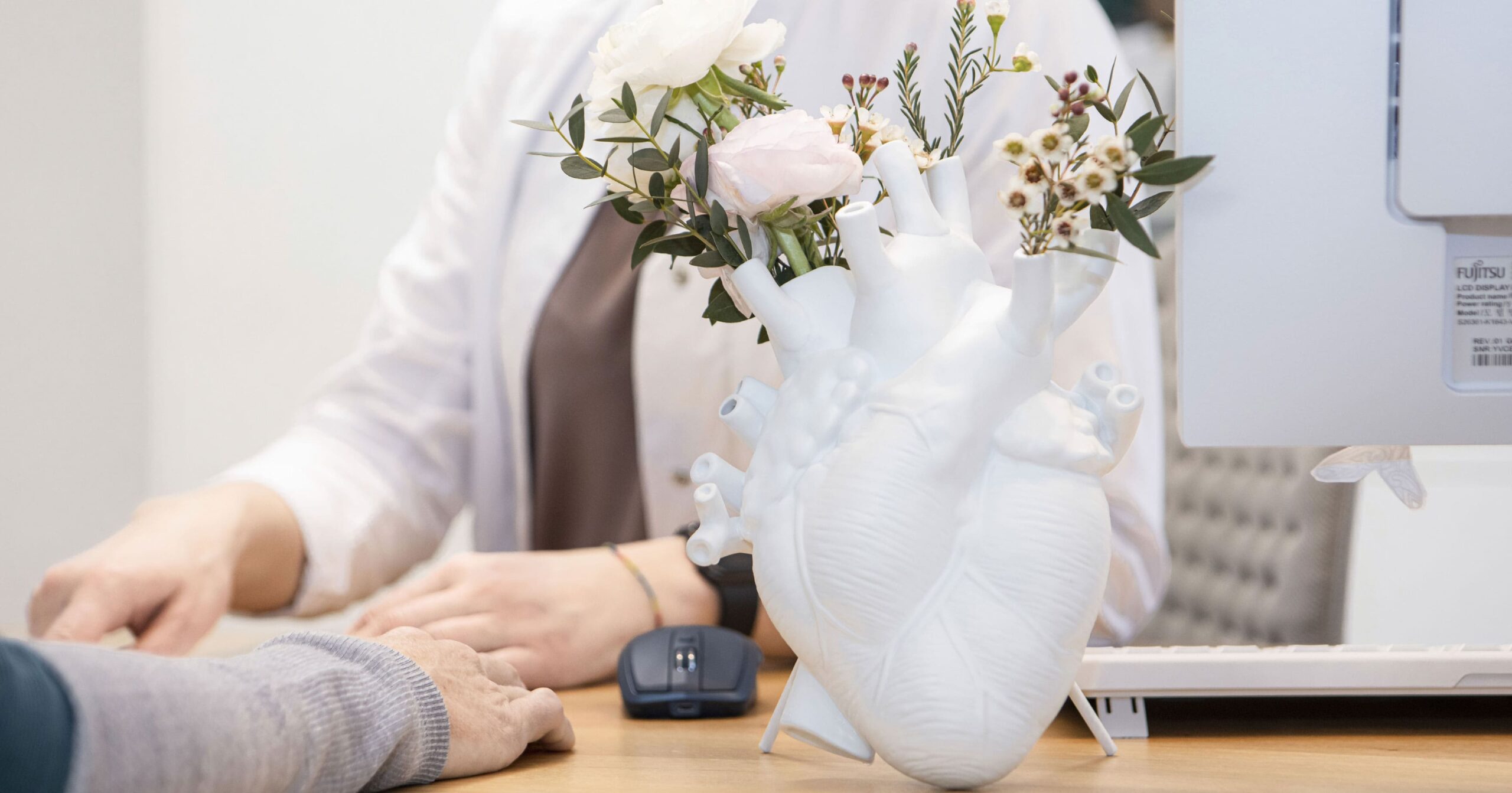 An einem Schreibtisch sieht man eine weiße Blumenvase, die die Form eines anatomischen Herzens hat und in der eine gelbe und eine rosa Rose sowie weitere Pflanzen stecken, im Hintergrund sitzen Dr. Dissauer und Patient am Tisch