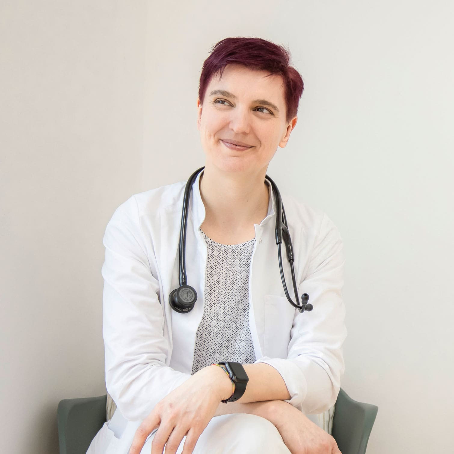 Dr. Dissauer, eine Dame mit rötlich gefärbten, kurzen Haaren, sitzt im Arztkittel und Stethoskop um den Hals auf einem Stuhl, blickt nach rechts lächelt