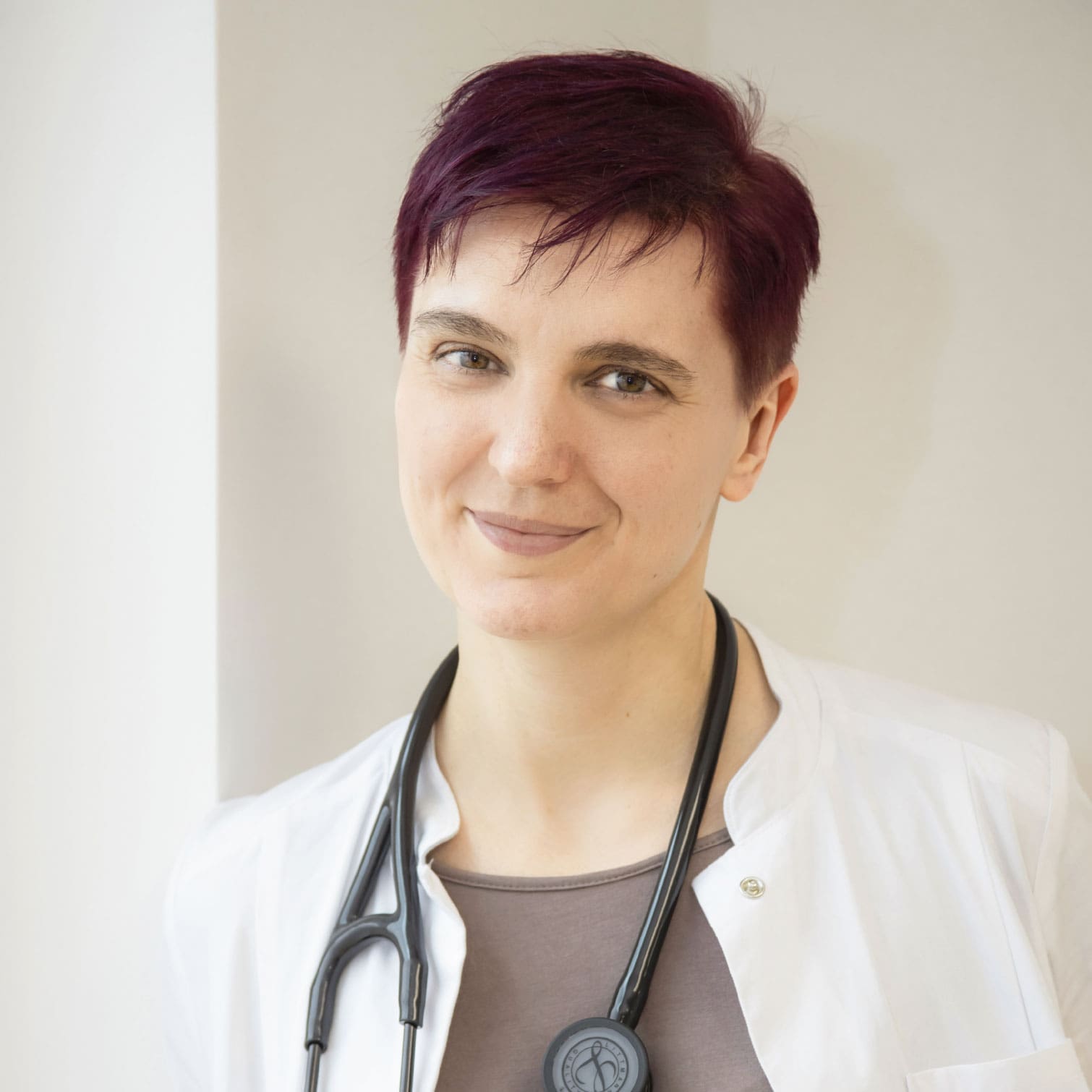 Dr. Dissauer, eine Dame mit rötlich gefärbten, kurzen Haaren, blickt im Arztkittel und Stethoskop um den Hals lächelnd in die Kamera