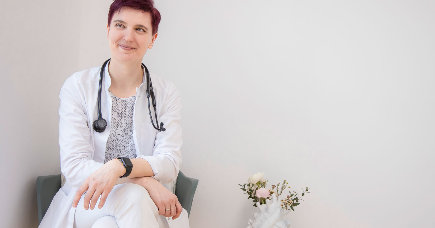 Dr. Dissauer, eine Dame mit rötlich gefärbten, kurzen Haaren, sitzt im Arztkittel und Stethoskop um den Hals auf einem Stuhl, blickt nach rechts und lächelt