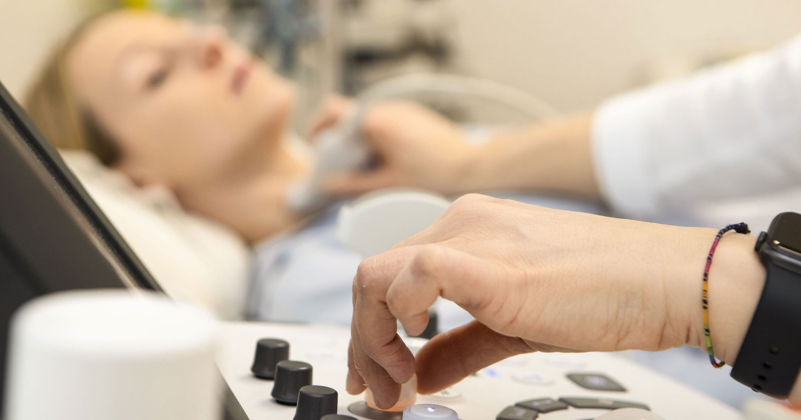 Nahaufnahme eines Ultraschalluntersuchungsgerätes, im Hintergrund eine Frau, deren Schilddrüse untersucht wird