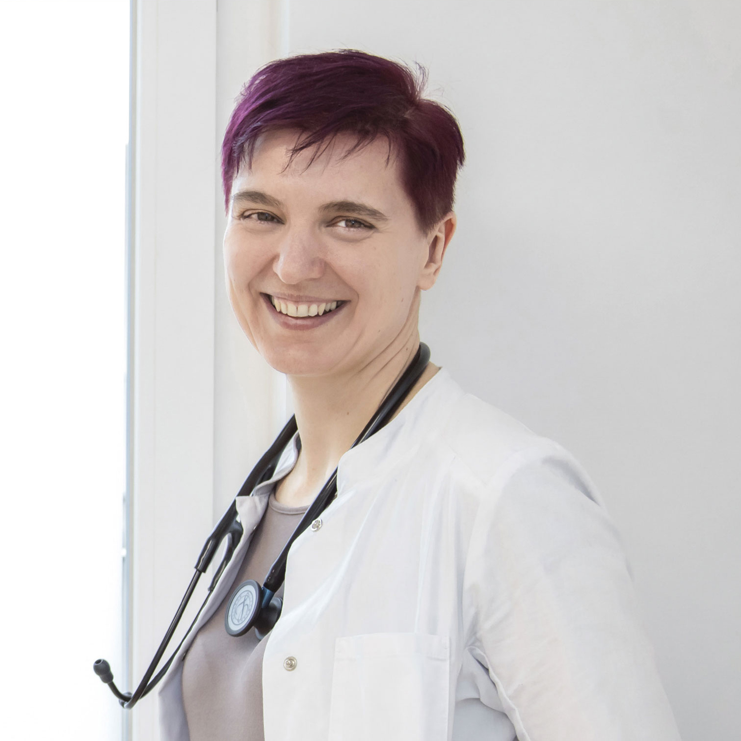 Dr. Dissauer, eine Dame mit rötlich gefärbten, kurzen Haaren, blickt im Arztkittel und Stethoskop um den Hals von der Seite lächelnd in die Kamera